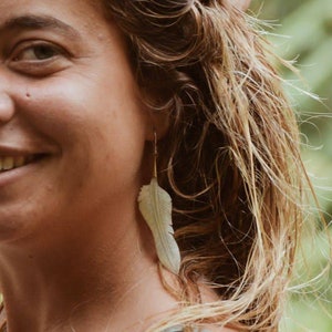 Op de afbeelding: Een vrouw met lang, nat, blond haar glimlacht en kijkt naar de camera. Ze draagt een lichtgrijs tanktop en heeft een grote, witte veren oorbel in haar linker oor.