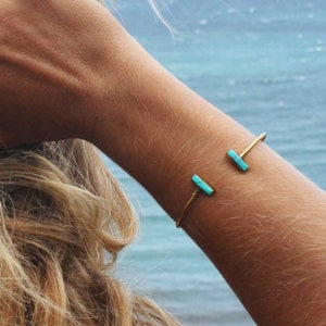 Può includere: Un braccialetto d'oro con due pietre turchesi sul polso di una donna. Il braccialetto è aperto e ha un design semplice.