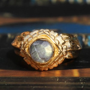 Puede incluir: Un anillo de oro con una gran piedra preciosa gris facetada engastada en un bisel cuadrado. El anillo tiene un diseño detallado y texturizado.