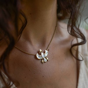 Puede incluir: Un collar de cadena dorada con un colgante blanco y dorado en forma de pájaro con las alas extendidas.