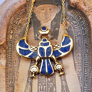 Puede incluir: Un collar dorado con un colgante que presenta un diseño de escarabajo azul y dorado. El collar se muestra sobre un fondo de una ilustración de un sarcófago egipcio antiguo.