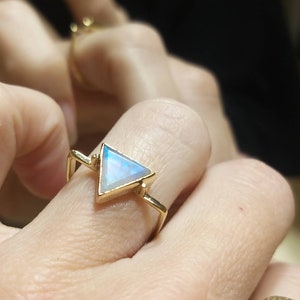 May include: A gold ring featuring a triangular iridescent gemstone. The ring is worn on a finger.