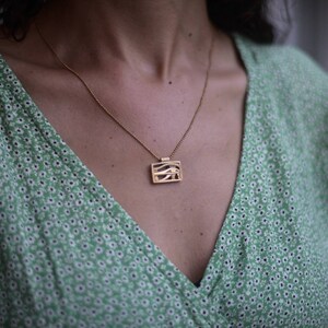 May include: A gold chain necklace with a rectangular pendant featuring a stylized eye design. The necklace is worn by a person wearing a green floral patterned dress.