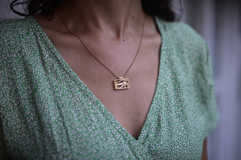 May include: A gold chain necklace with a rectangular pendant featuring a stylized eye design. The necklace is worn by a person wearing a green floral patterned dress.