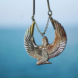 May include: A gold chain necklace with a pendant featuring a detailed depiction of a winged goddess with outstretched wings. The pendant is made of a light brown material with intricate details.