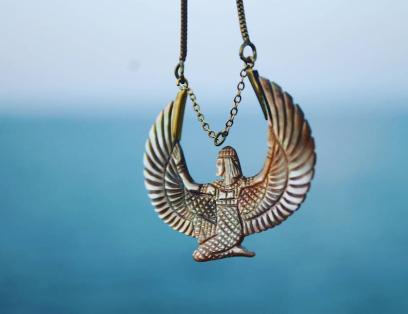 May include: A gold chain necklace with a pendant featuring a detailed depiction of a winged goddess with outstretched wings. The pendant is made of a light brown material with intricate details.