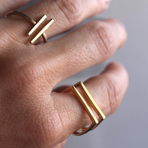 Puede incluir: Primer plano de dos anillos dorados con un diseño minimalista. Un anillo presenta dos barras paralelas, mientras que el otro tiene forma rectangular con dos barras paralelas. Los anillos se llevan en una mano, mostrando su estilo simple pero elegante.
