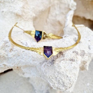 Puede incluir: Dos pulseras doradas con gemas facetadas en forma de escudo. Las gemas presentan un degradado de tonos morados y azules. Las pulseras están sobre una superficie texturizada de color blanquecino. Joyería.