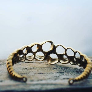 Puede incluir: Un brazalete de metal dorado con un diseño único y calado que presenta una serie de círculos interconectados. El brazalete está sobre una superficie de madera.