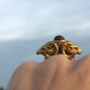 Op de afbeelding: Een ring van goudkleurige metaal met drie rammenkoppen en een donkerpaarse steen in het midden.