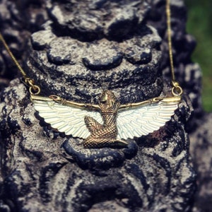 Puede incluir: Un collar de oro con un colgante que representa una figura alada con una corona. El colgante est&aacute; hecho de metal y tiene un acabado blanco iridiscente.
