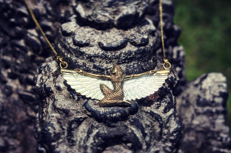 Puede incluir: Un collar de oro con un colgante que representa una figura alada con una corona. El colgante est&aacute; hecho de metal y tiene un acabado blanco iridiscente.