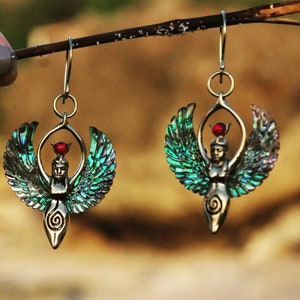 Puede incluir: Un par de pendientes con una figura de diosa alada con una corona de piedras preciosas rojas. Las alas están hechas de concha de abulón verde y azul iridiscente.