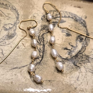 Puede incluir: Un par de pendientes colgantes dorados con perlas blancas. Los pendientes se exhiben sobre una superficie decorativa con una ilustración vintage. Las perlas están ensartadas con un delicado alambre dorado.