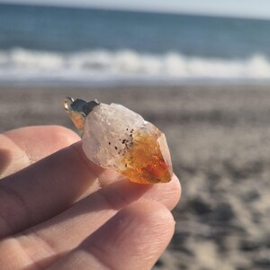 Puede incluir: Un colgante de cristal de citrino con una fianza dorada. El cristal es una mezcla de tonos claros y ámbar, con una punta. El fondo muestra una playa de arena y olas del océano.
