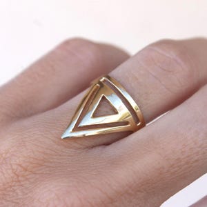 May include: A gold-coloured ring with a geometric triangle design. The ring features a hollow triangle shape within a larger triangle, creating a modern and minimalist aesthetic. The ring is worn on a finger.