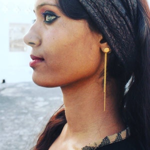 May include: A woman wearing a black headscarf with gold chain earrings. The earrings have a small round disc at the top and a long chain that hangs down to her neck.