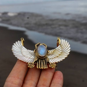 May include: A gold pendant necklace featuring a scarab beetle design with an iridescent moonstone. The wings are crafted from white mother of pearl.