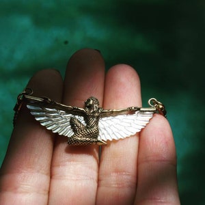 Puede incluir: Un collar de oro con un colgante que representa una figura alada con un dise&ntilde;o blanco y dorado.
