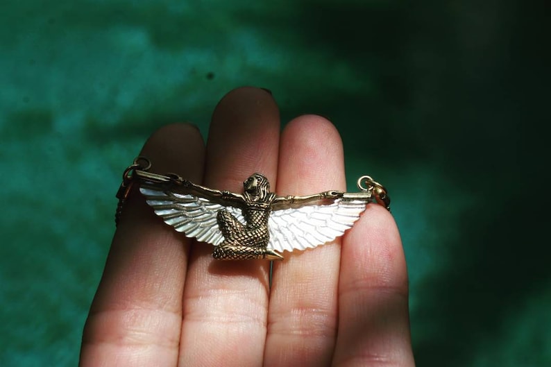 Puede incluir: Un collar de oro con un colgante que representa una figura alada con un dise&ntilde;o blanco y dorado.