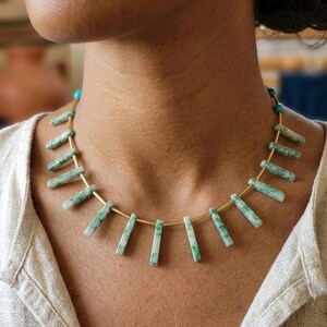 Puede incluir: Un collar con una base de alambre metálico fino en tono dorado adornado con múltiples cuentas rectangulares colgantes de piedra veteada en verde y blanco. Pequeñas cuentas esféricas de color turquesa se sitúan en los extremos del alambre.
