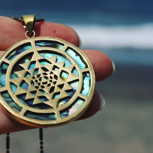 Puede incluir: Colgante redondo dorado con un intrincado diseño geométrico. El colgante presenta un patrón central de triángulos entrelazados y está incrustado con concha de abulón iridiscente azul y verde. Un pequeño lazo en la parte superior para una cadena.