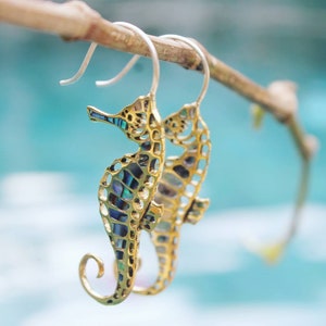 Puede incluir: Un par de pendientes de tono dorado con un diseño de trabajo abierto detallado de un caballito de mar. Los caballitos de mar están adornados con acentos de concha de abulón iridiscente.