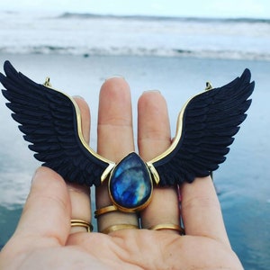 May include: A gold necklace featuring black wings and a teardrop-shaped blue gemstone pendant.