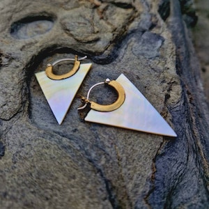 May include: A pair of gold triangle earrings with a mother-of-pearl inlay. The earrings are hanging on a rough, textured surface.
