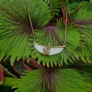 Puede incluir: Un collar de cadena dorada con un colgante blanco y dorado que representa una figura alada. El colgante tiene la forma de una mujer con las alas extendidas.