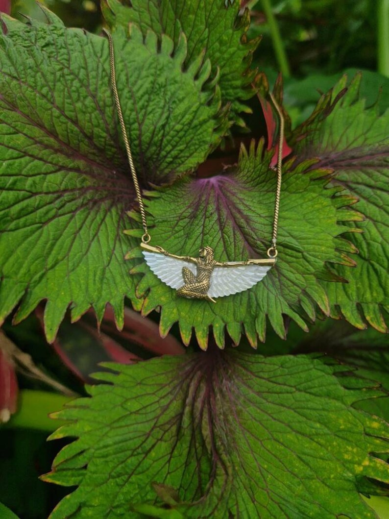 Puede incluir: Un collar de cadena dorada con un colgante blanco y dorado que representa una figura alada. El colgante tiene la forma de una mujer con las alas extendidas.