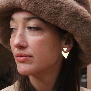 Puede incluir: Una mujer con un sombrero de piel sintética marrón con pendientes triangulares dorados. También lleva un aro de nariz dorado.