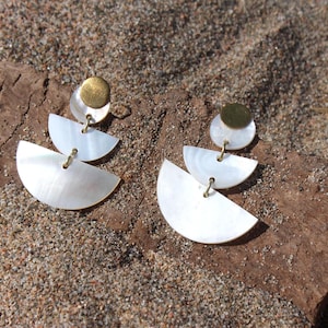 Puede incluir: Un par de pendientes elegantes con un disco de latón y tres medias lunas de nácar. Los pendientes están conectados con pequeños anillos dorados. Los pendientes se exhiben sobre un trozo de madera flotante.