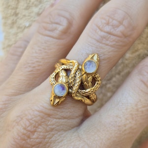 Puede incluir: Un anillo dorado con un diseño de serpiente. El anillo tiene dos cabezas de serpiente enfrentadas, cada una con una piedra redonda e iridiscente. Los cuerpos de las serpientes están entrelazados, creando una joya única y detallada.