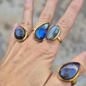 Puede incluir: Cuatro anillos dorados con grandes piedras de labradorita azul iridiscente se muestran en dedos. Las piedras tienen varias formas, incluyendo ovaladas y de lágrima. Los anillos tienen un engaste sencillo y elegante.