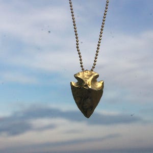 Puede incluir: Un colgante de punta de flecha de color dorado cuelga de un collar de cadena de bolas. La punta de flecha tiene una superficie texturizada y una forma triangular, con una base dentada. El fondo es un cielo nublado.