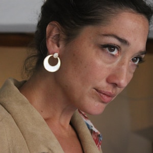 Puede incluir: Una mujer con el pelo oscuro que lleva una chaqueta marrón y un par de pendientes de aro plateados con un pequeño detalle redondo.