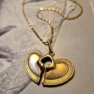 May include: A gold-colored necklace with a pendant featuring a broken heart design. The pendant has a key-shaped element that fits into the heart. The chain is delicate and gold-toned.