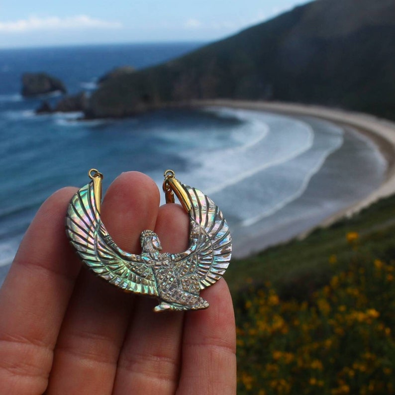 Puede incluir: Un collar con un colgante dorado e iridiscente que presenta un dise&ntilde;o detallado de una diosa alada con las alas extendidas. El colgante se sostiene en una mano sobre un fondo de playa y oc&eacute;ano.
