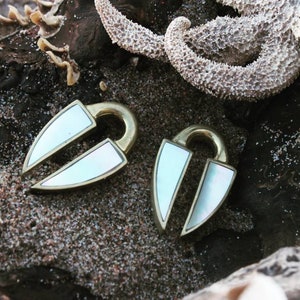 May include: A pair of brass ear gauges with a white mother-of-pearl inlay. The gauges are shaped like a heart with a split in the middle. The gauges are on a sandy surface.