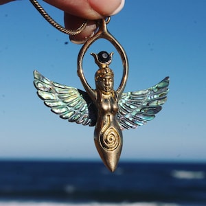 Puede incluir: Un collar con un colgante de oro que representa a una diosa alada con una piedra preciosa negra en la parte superior de su cabeza. Las alas están hechas de concha de abulón iridiscente.