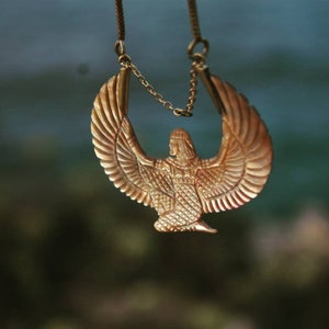 May include: A gold necklace with a pendant featuring a winged figure with a crown. The pendant is detailed with intricate patterns and a textured surface.