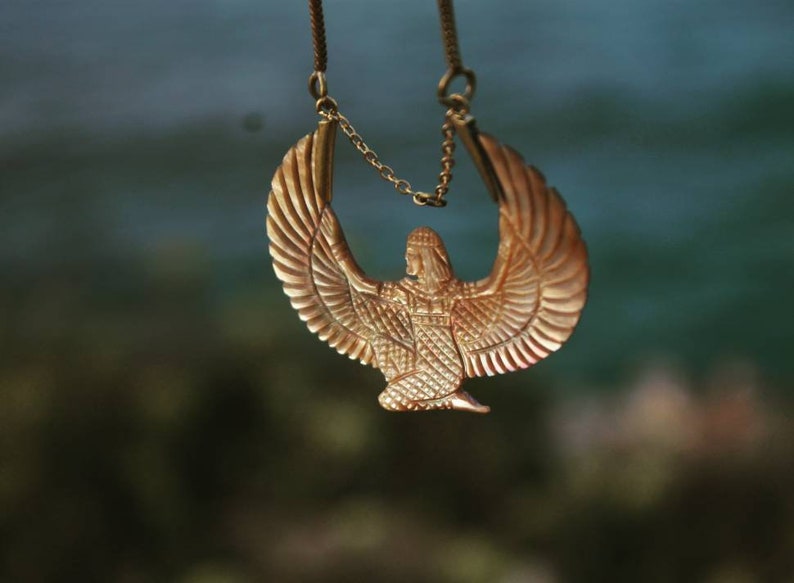 May include: A gold necklace with a pendant featuring a winged figure with a crown. The pendant is detailed with intricate patterns and a textured surface.