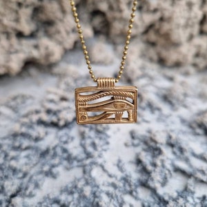 May include: A gold chain necklace with a square pendant featuring an intricate design of the Eye of Horus. The pendant is made of metal and has a detailed, textured surface.