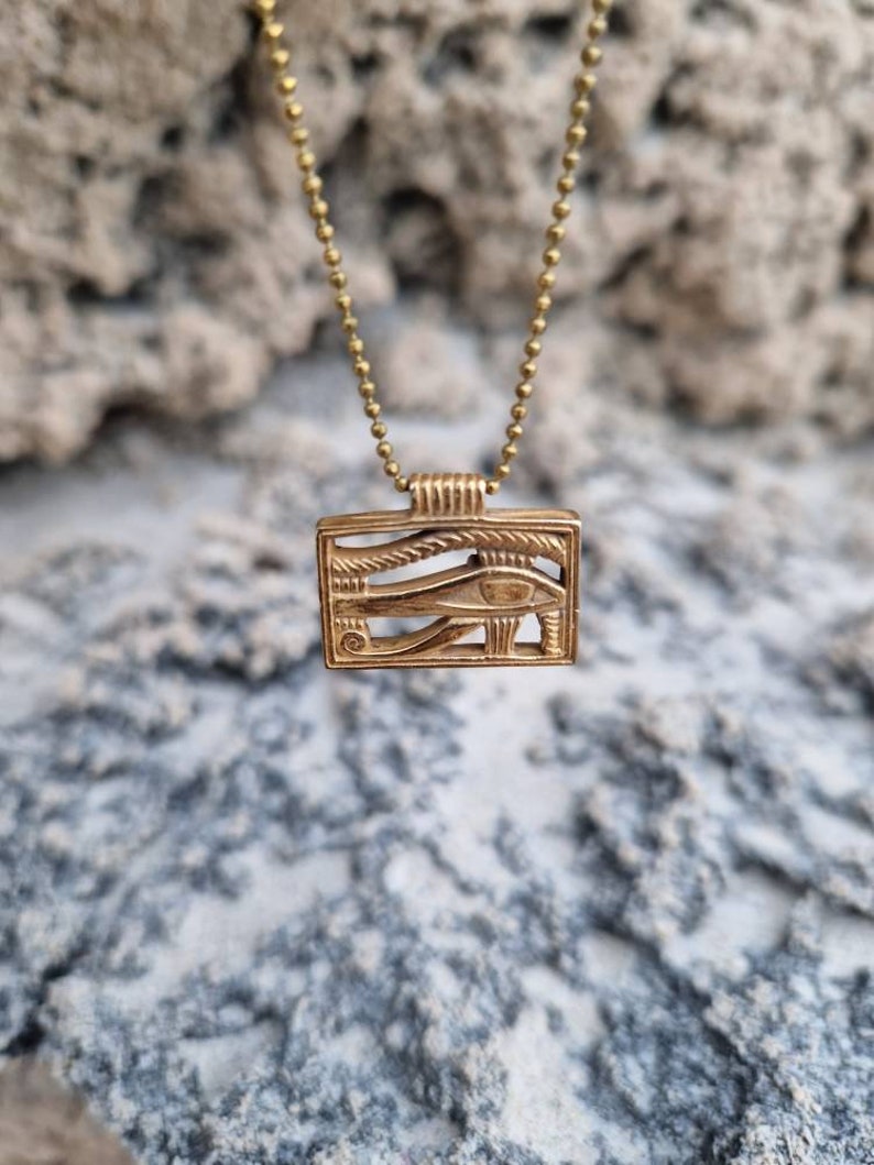 May include: A gold chain necklace with a square pendant featuring an intricate design of the Eye of Horus. The pendant is made of metal and has a detailed, textured surface.