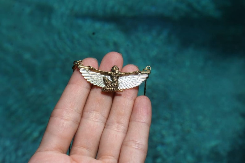 Puede incluir: Un collar de oro con un colgante que representa una figura alada con alas blancas. El colgante se sostiene en una mano sobre un fondo azul.
