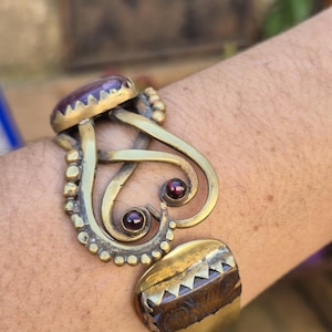 Puede incluir: Una pulsera de puño de latón con una gran piedra ovalada de amatista y dos piedras rojas más pequeñas. La pulsera tiene un diseño ornamental y arremolinado con un cierre rectangular.