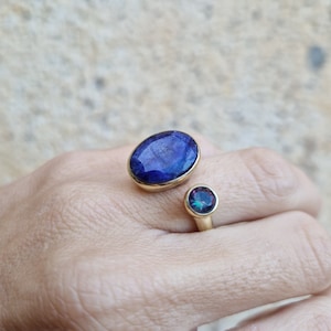 Könnte beinhalten: Ein goldener Ring mit zwei blauen Edelsteinen. Der größere Edelstein ist oval geformt und der kleinere Edelstein ist rund. Der Ring befindet sich an einem Finger.