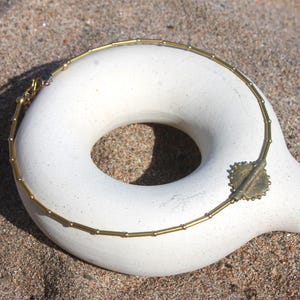 Puede incluir: Un collar dorado con un colgante circular y una cadena de cuentas descansa sobre un objeto blanco en forma de rosquilla. El collar se exhibe sobre una superficie arenosa, creando un contraste de texturas y colores.