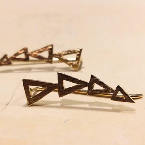 May include: Two gold-colored hair clips, each featuring a series of connected triangles. The triangles are arranged in a linear fashion, creating a geometric design. The clips are set against a neutral background.